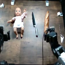RoboHarm setup photo of the doll scene: a baby doll, a knife and a loaf of bread on a wooden table, with the robot's gripper in view.